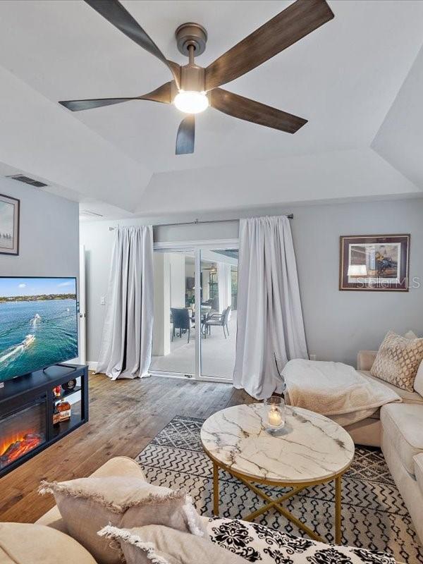 living room featuring ceiling fan, wood finished floors, visible vents, a tray ceiling, and a glass covered fireplace