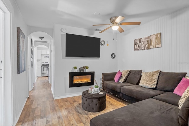 living area featuring a warm lit fireplace, arched walkways, baseboards, ceiling fan, and light wood-type flooring