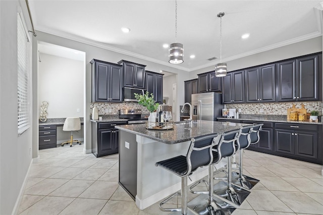 kitchen featuring light tile patterned floors, built in study area, an island with sink, appliances with stainless steel finishes, and a breakfast bar