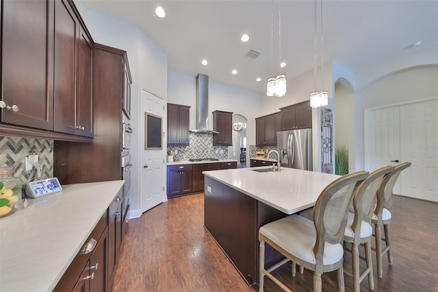 kitchen with pendant lighting, a center island with sink, stainless steel appliances, light countertops, and wall chimney range hood