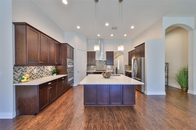 kitchen featuring arched walkways, stainless steel appliances, hanging light fixtures, light countertops, and wall chimney exhaust hood