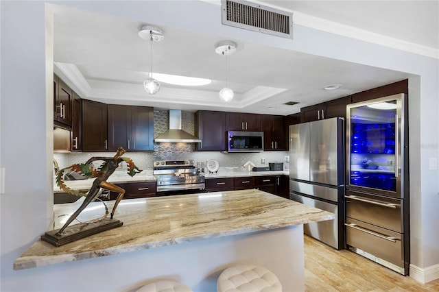 kitchen featuring stainless steel appliances, a raised ceiling, visible vents, a peninsula, and wall chimney exhaust hood