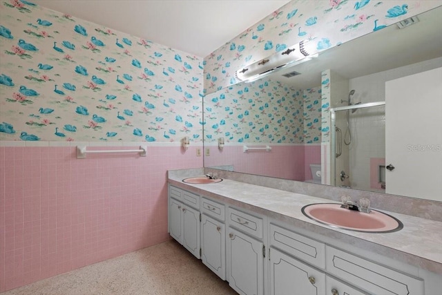 bathroom featuring double vanity, a stall shower, wainscoting, a sink, and wallpapered walls