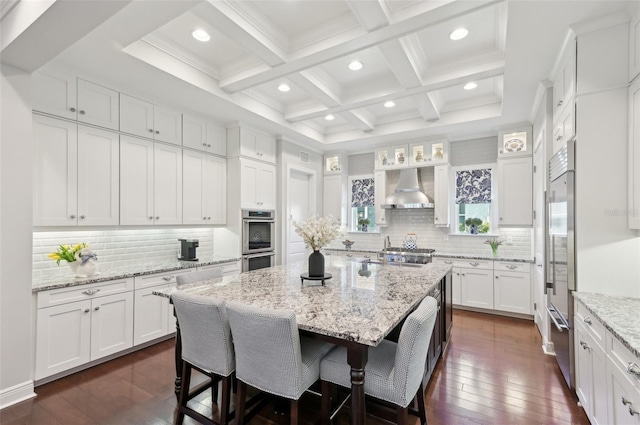 kitchen featuring coffered ceiling, wall chimney range hood, a kitchen bar, white cabinetry, and beam ceiling