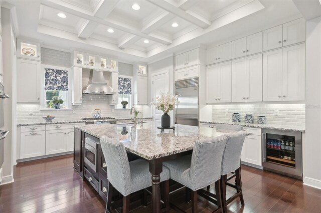 kitchen featuring built in appliances, wine cooler, a breakfast bar area, white cabinetry, and wall chimney range hood