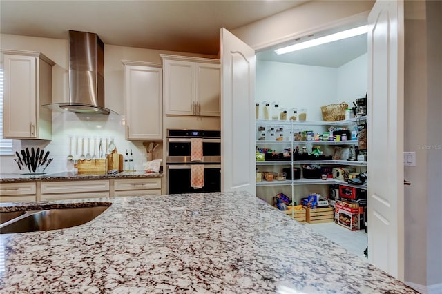 kitchen with light stone counters, a sink, backsplash, double oven, and wall chimney range hood