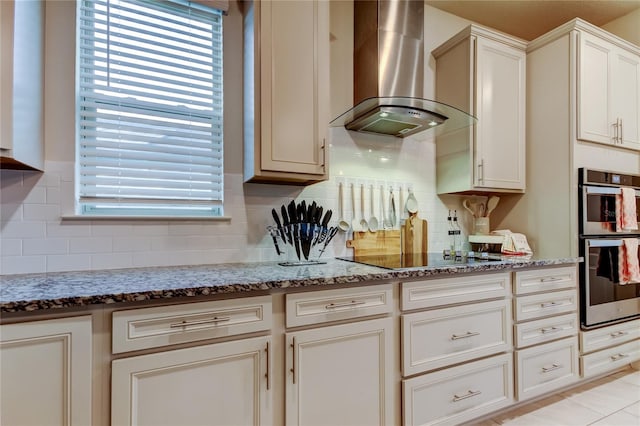 kitchen featuring light stone counters, stainless steel double oven, black electric stovetop, wall chimney exhaust hood, and tasteful backsplash