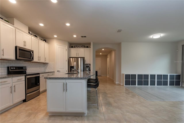 kitchen featuring arched walkways, stone counters, a kitchen island with sink, white cabinetry, and appliances with stainless steel finishes