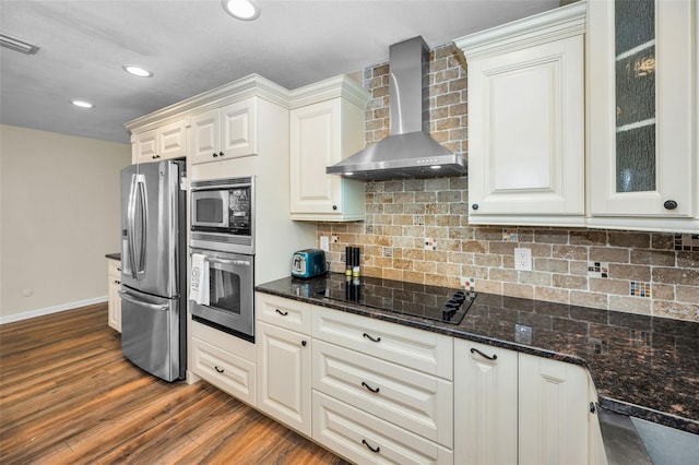 kitchen featuring stainless steel appliances, dark stone counters, glass insert cabinets, and wall chimney range hood