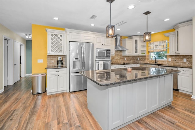 kitchen with appliances with stainless steel finishes, wall chimney range hood, a center island, glass insert cabinets, and pendant lighting