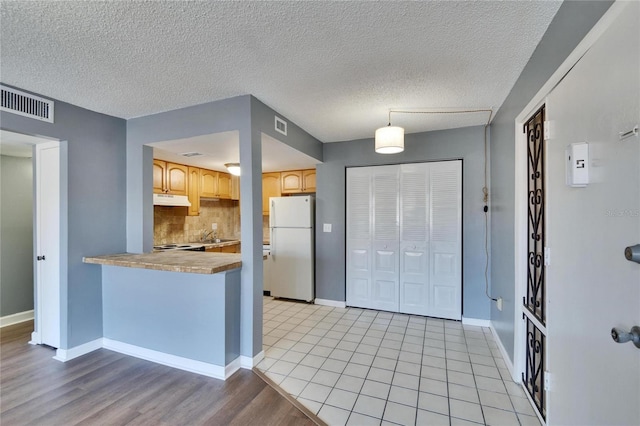 kitchen with tasteful backsplash, visible vents, freestanding refrigerator, light brown cabinets, and under cabinet range hood