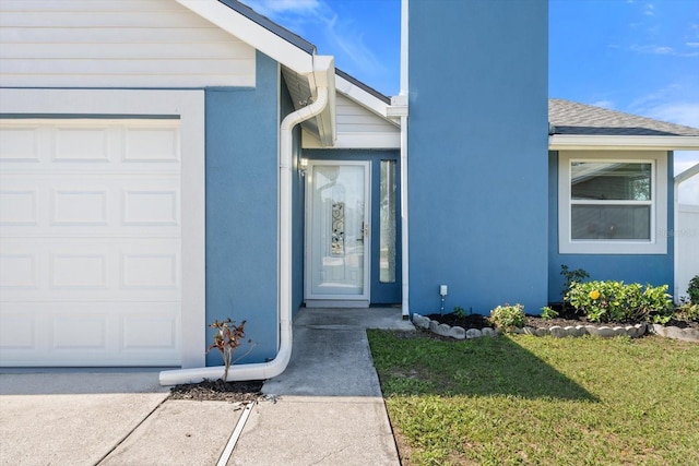 view of exterior entry with a garage, a yard, a shingled roof, and stucco siding