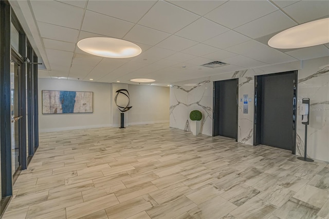 finished basement featuring a paneled ceiling, visible vents, and elevator