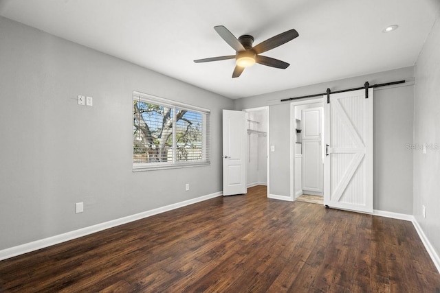 unfurnished bedroom featuring ceiling fan, a barn door, dark wood-style flooring, and baseboards