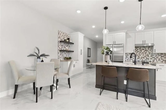 kitchen featuring freestanding refrigerator, a kitchen island with sink, under cabinet range hood, open shelves, and backsplash