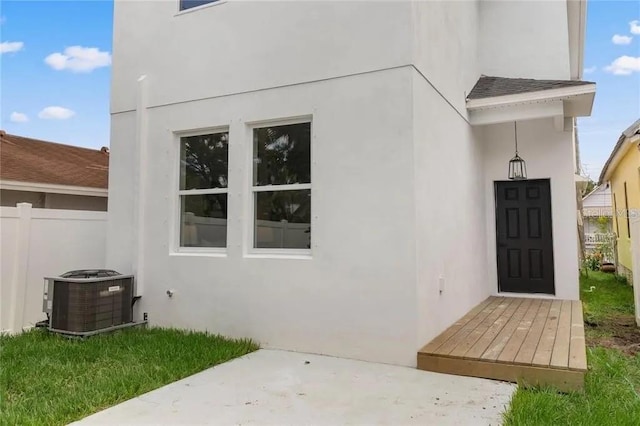 entrance to property with central AC unit, roof with shingles, fence, and stucco siding