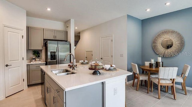 kitchen featuring stainless steel fridge, an island with sink, light countertops, gray cabinetry, and a sink