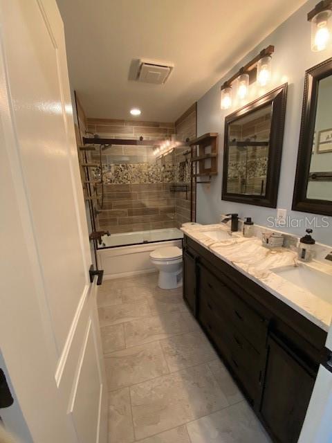 full bathroom with double vanity, visible vents, bath / shower combo with glass door, toilet, and a sink