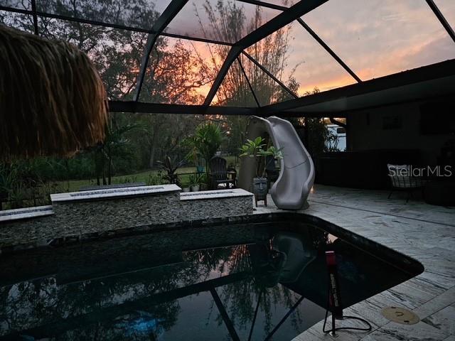 pool at dusk with a hot tub, glass enclosure, a patio area, a water slide, and an outdoor pool