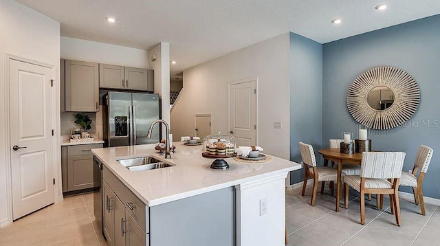 kitchen with gray cabinets, light countertops, a sink, an island with sink, and stainless steel fridge