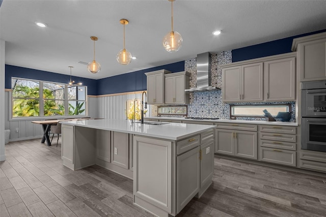 kitchen with a center island with sink, light countertops, hanging light fixtures, a sink, and wall chimney exhaust hood