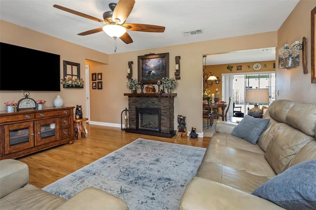 living room with a stone fireplace, wood finished floors, a ceiling fan, visible vents, and baseboards