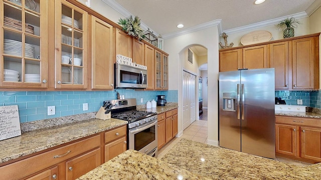 kitchen with arched walkways, light stone counters, appliances with stainless steel finishes, ornamental molding, and glass insert cabinets