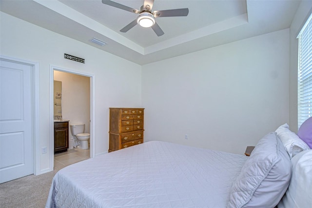 bedroom with a tray ceiling, light colored carpet, visible vents, a ceiling fan, and connected bathroom