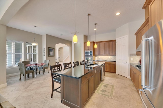 kitchen featuring stainless steel appliances, brown cabinetry, dark countertops, decorative light fixtures, and an island with sink