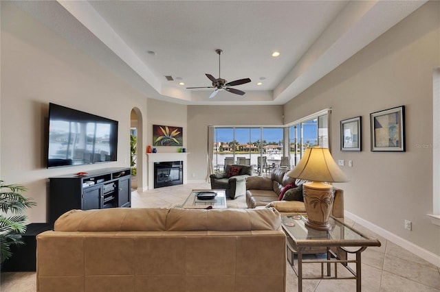 living room featuring light tile patterned floors, recessed lighting, baseboards, a tray ceiling, and a glass covered fireplace