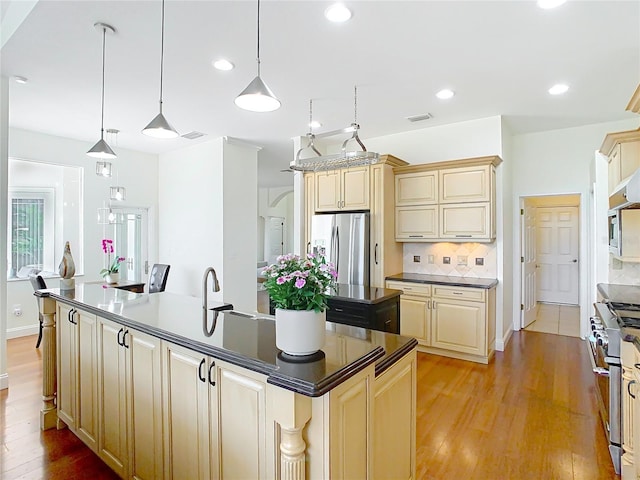 kitchen with cream cabinetry, an island with sink, dark countertops, stainless steel appliances, and decorative backsplash