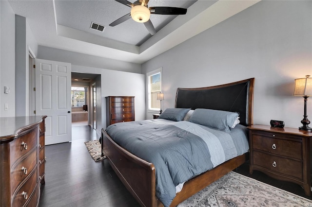 bedroom with visible vents, a tray ceiling, a textured ceiling, ceiling fan, and dark wood-style flooring