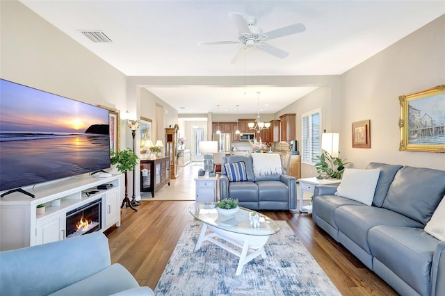 living area featuring ceiling fan with notable chandelier, hardwood / wood-style flooring, a glass covered fireplace, and visible vents