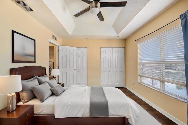 bedroom with dark wood-type flooring, two closets, visible vents, baseboards, and a raised ceiling