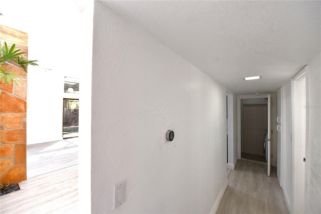 hall featuring a textured ceiling, a textured wall, light wood-style flooring, and baseboards