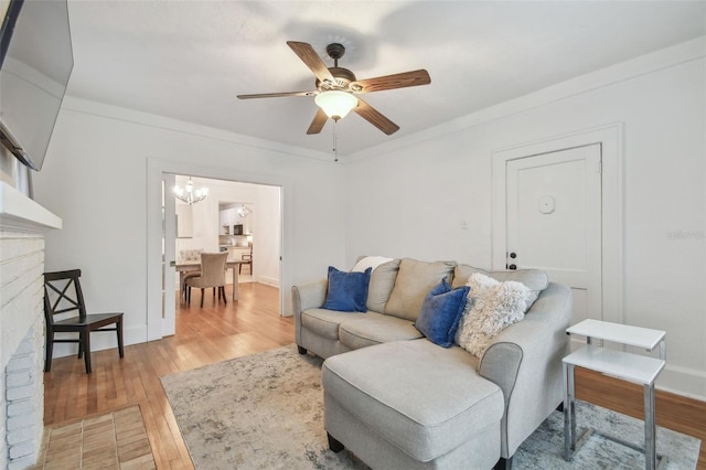 living area with a brick fireplace, ornamental molding, wood finished floors, and ceiling fan with notable chandelier