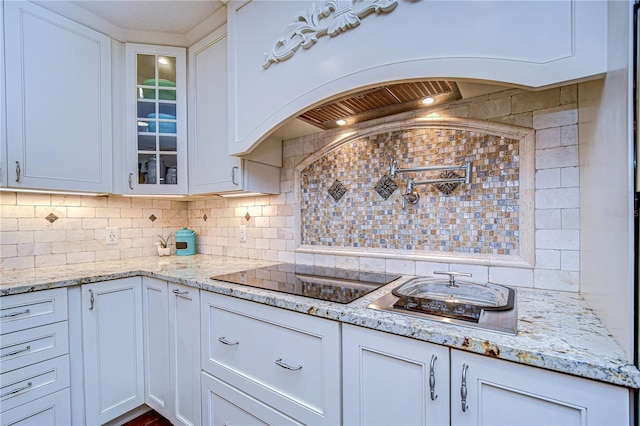 kitchen with black electric stovetop, white cabinetry, backsplash, light stone countertops, and glass insert cabinets