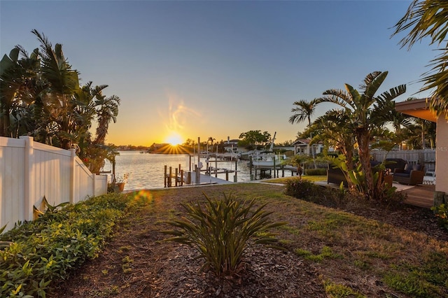 yard at dusk with a water view, boat lift, a boat dock, and fence