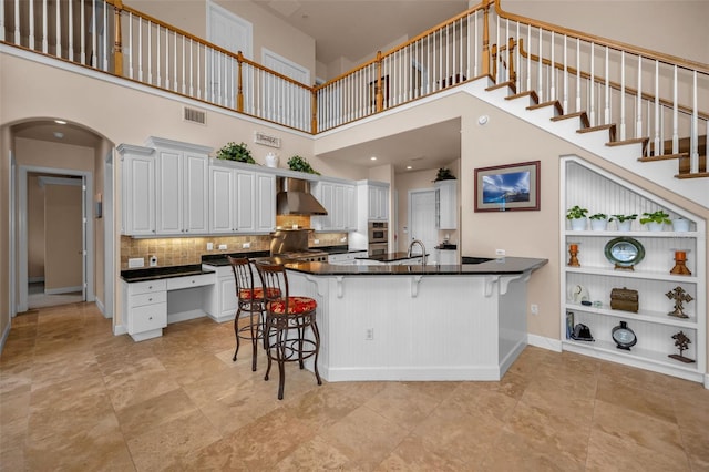 kitchen featuring visible vents, arched walkways, dark countertops, wall chimney exhaust hood, and a breakfast bar