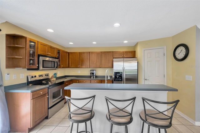 kitchen with light tile patterned floors, brown cabinetry, an island with sink, appliances with stainless steel finishes, and a breakfast bar area