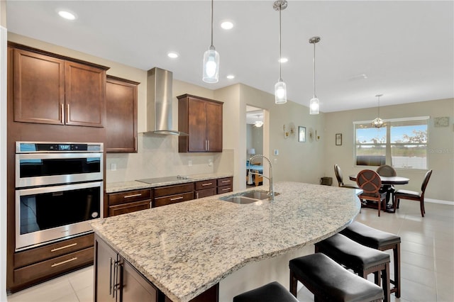 kitchen featuring pendant lighting, black electric stovetop, a sink, wall chimney range hood, and an island with sink