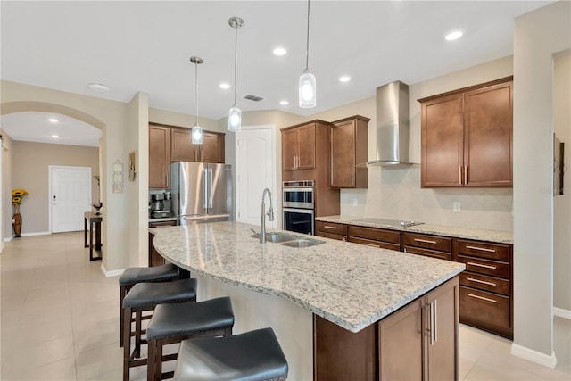 kitchen featuring a kitchen island with sink, stainless steel appliances, wall chimney range hood, light stone countertops, and pendant lighting