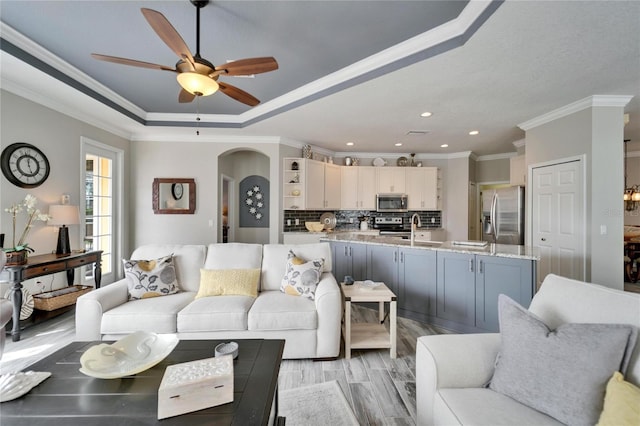 living room featuring arched walkways, ceiling fan, light wood-type flooring, a tray ceiling, and crown molding