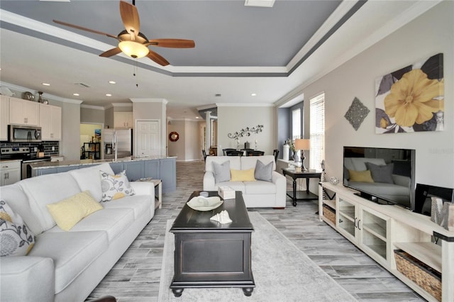 living room featuring ceiling fan, a tray ceiling, crown molding, light wood-style floors, and recessed lighting