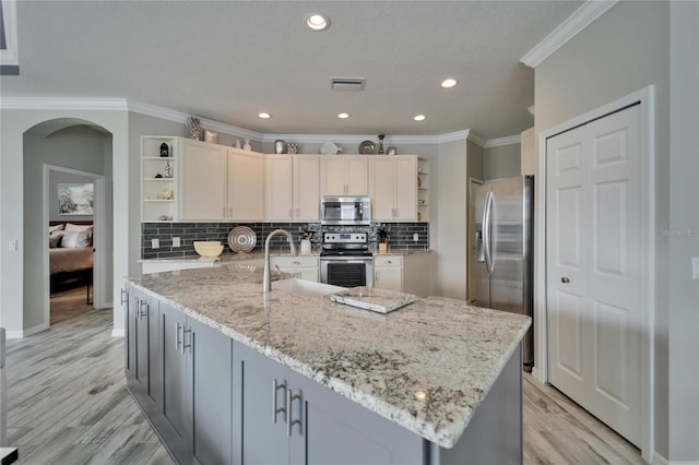 kitchen with arched walkways, light stone counters, a kitchen island with sink, stainless steel appliances, and open shelves