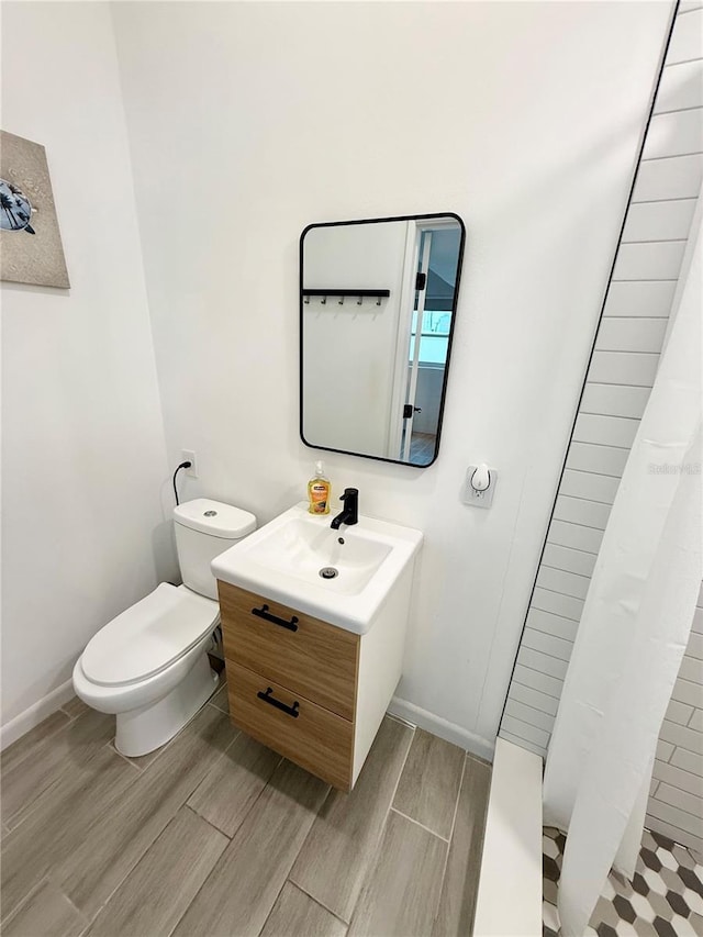 full bath featuring toilet, wood finish floors, vanity, baseboards, and a tile shower