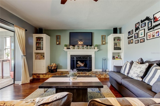 living room with baseboards, a fireplace with raised hearth, ceiling fan, dark wood-style flooring, and a textured ceiling