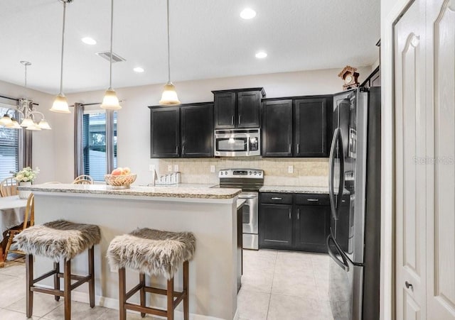 kitchen with light stone counters, a breakfast bar area, appliances with stainless steel finishes, dark cabinetry, and decorative light fixtures