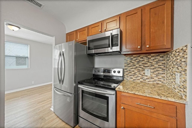 kitchen with brown cabinetry, light stone counters, stainless steel appliances, light wood-type flooring, and backsplash