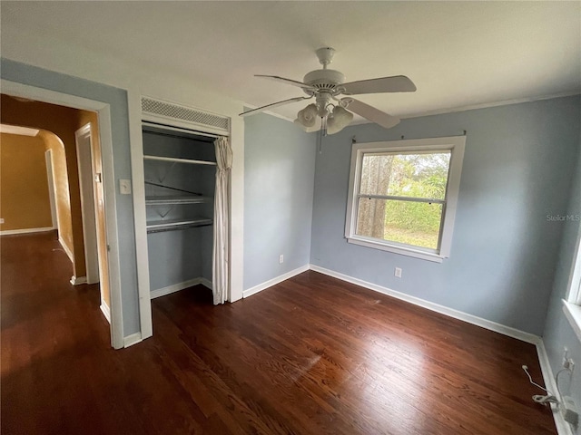 unfurnished bedroom featuring arched walkways, dark wood-style flooring, a ceiling fan, baseboards, and a closet